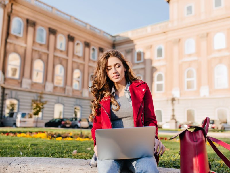 Outdoor portrait of serious curly female student sitting with laptop on the ground. Busy brunette girl in red jacket working with computer in front of college in warm day.